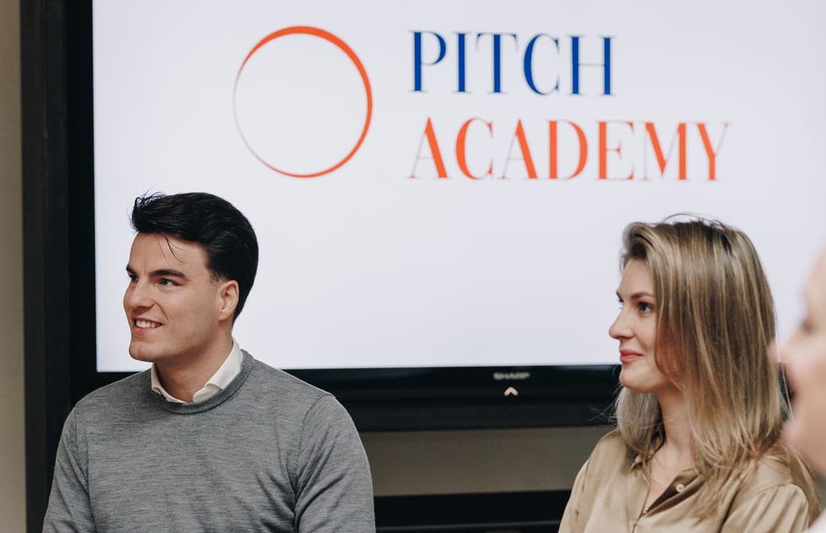 Deelnemers luisteren naar een pitch, met het logo van Pitch Academy op het scherm